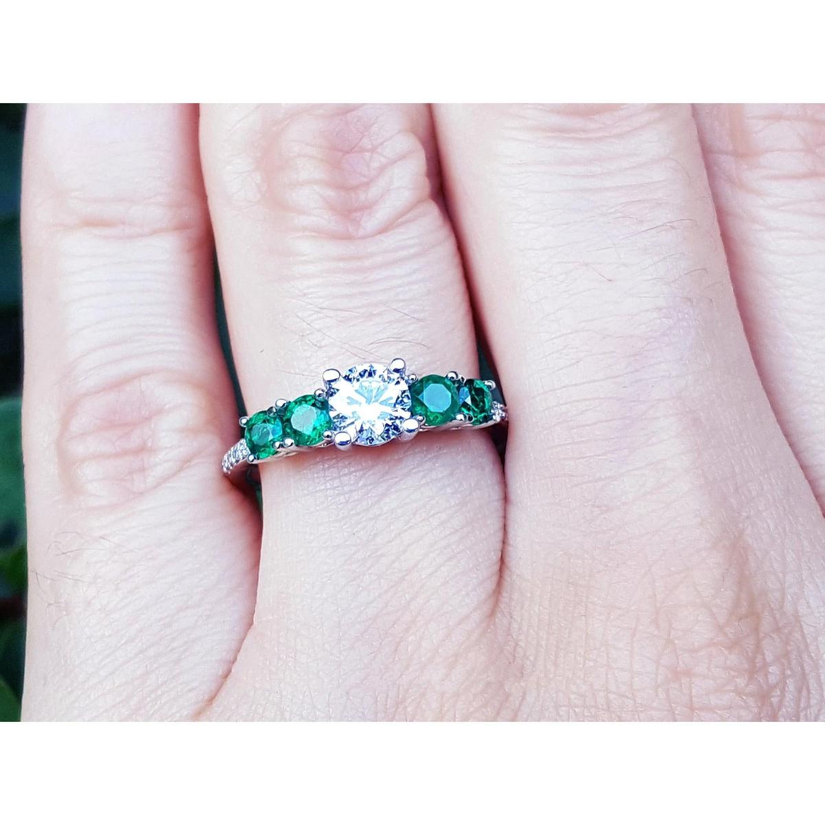 A ring with a center round diamond and side round emeralds on a finger.