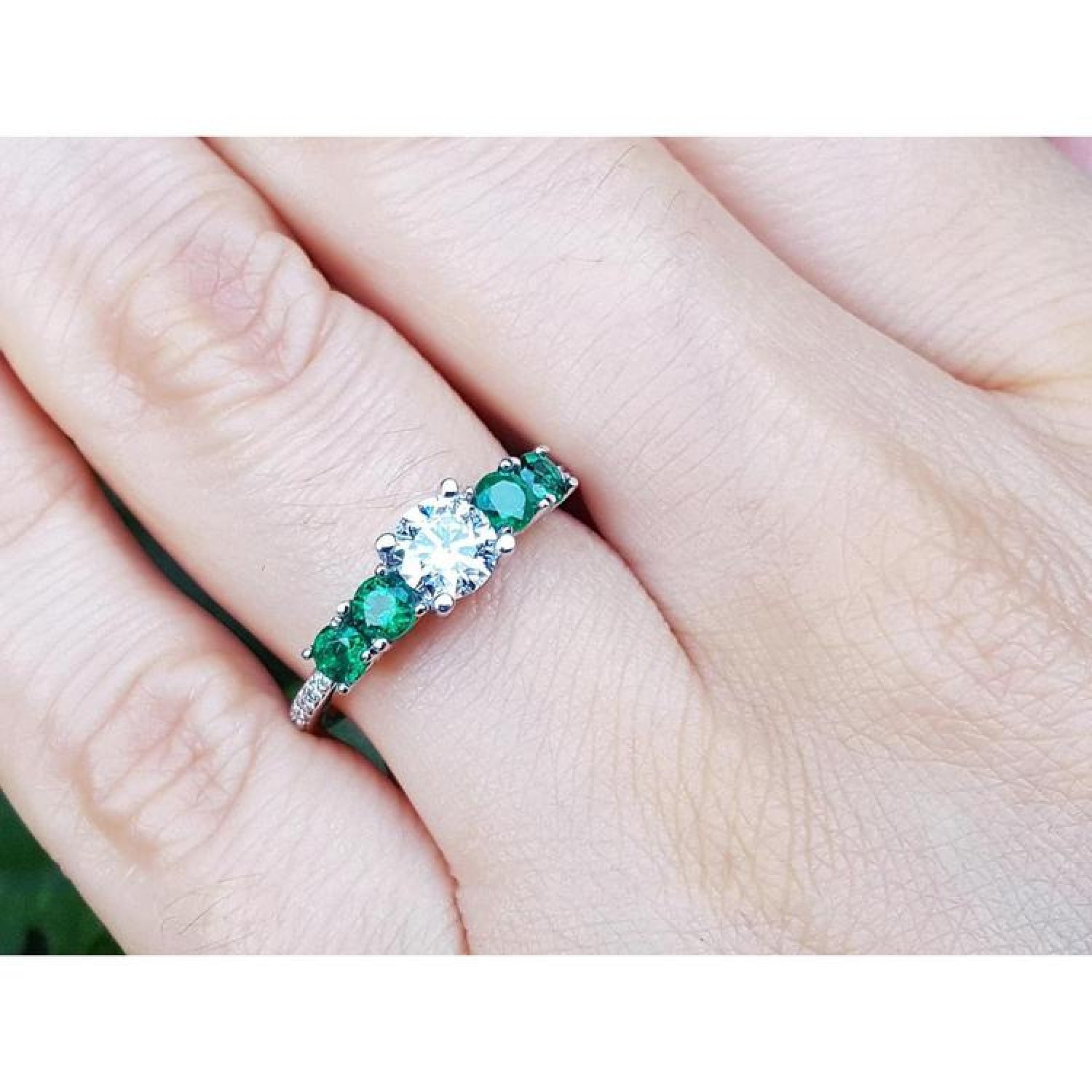 14K white gold ring with a round natural diamond and four side emeralds on a finger.