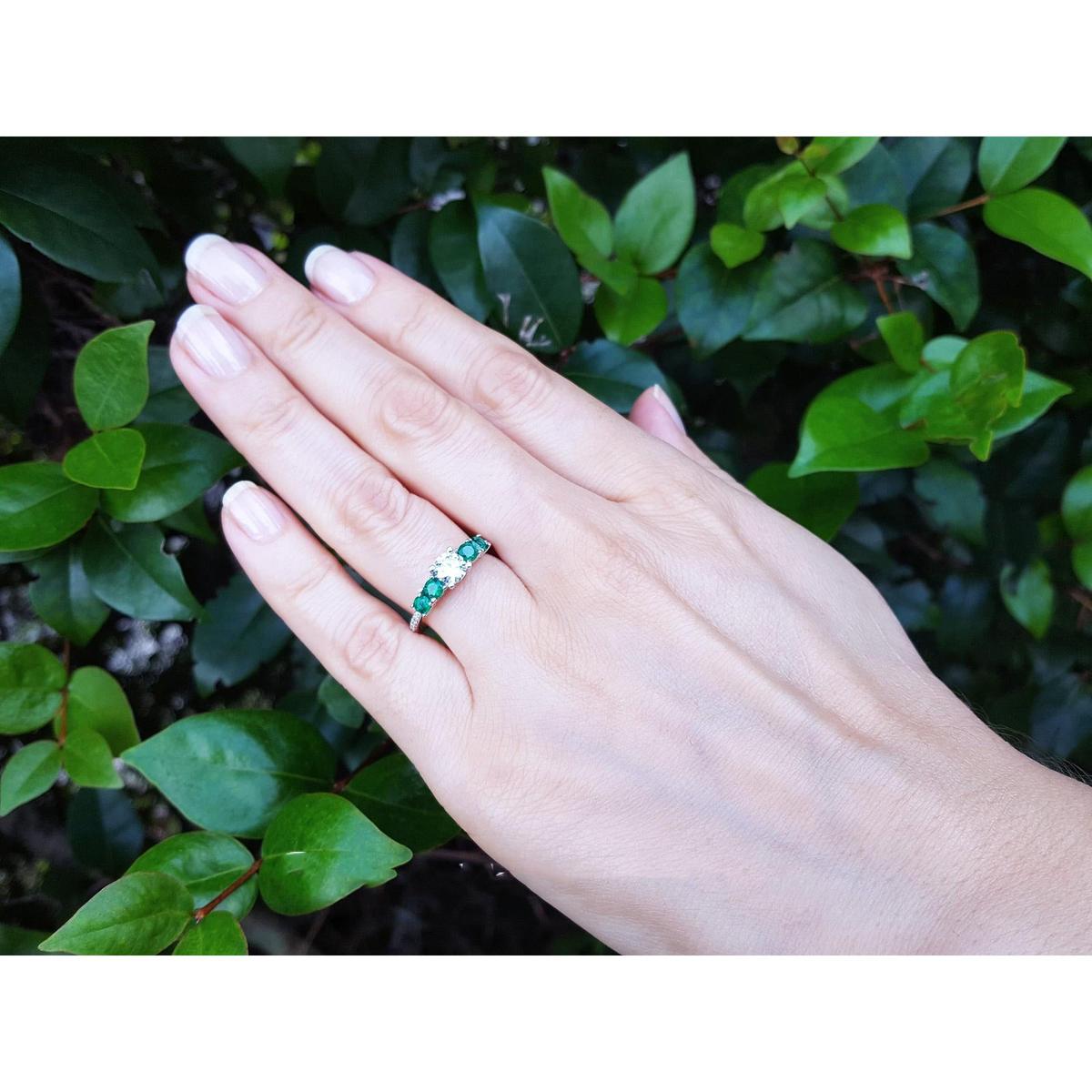 Hand wearing a ring with center diamond and side emeralds against a green leafy background.