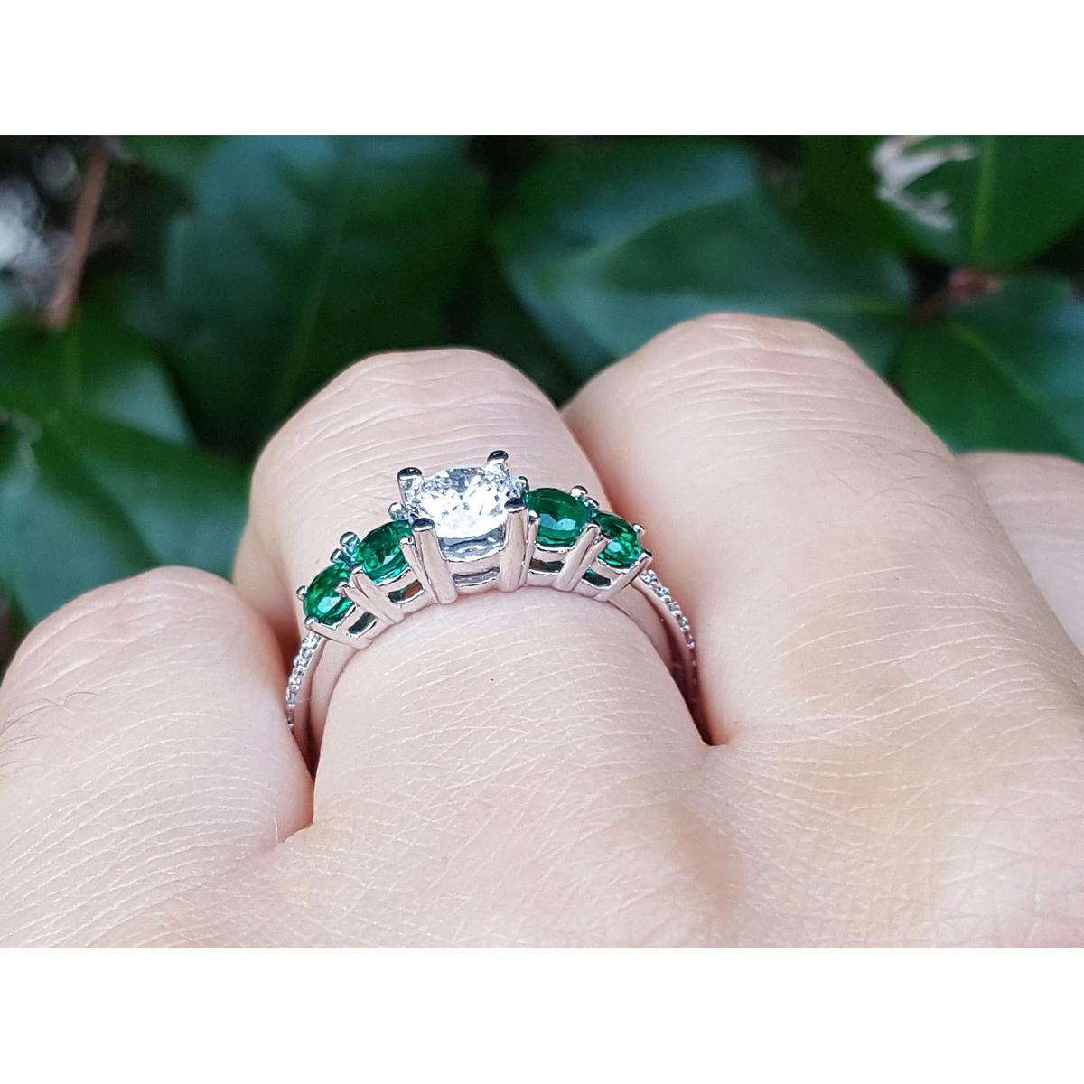 Ring with emeralds and a diamond on a finger against a green leafy background.