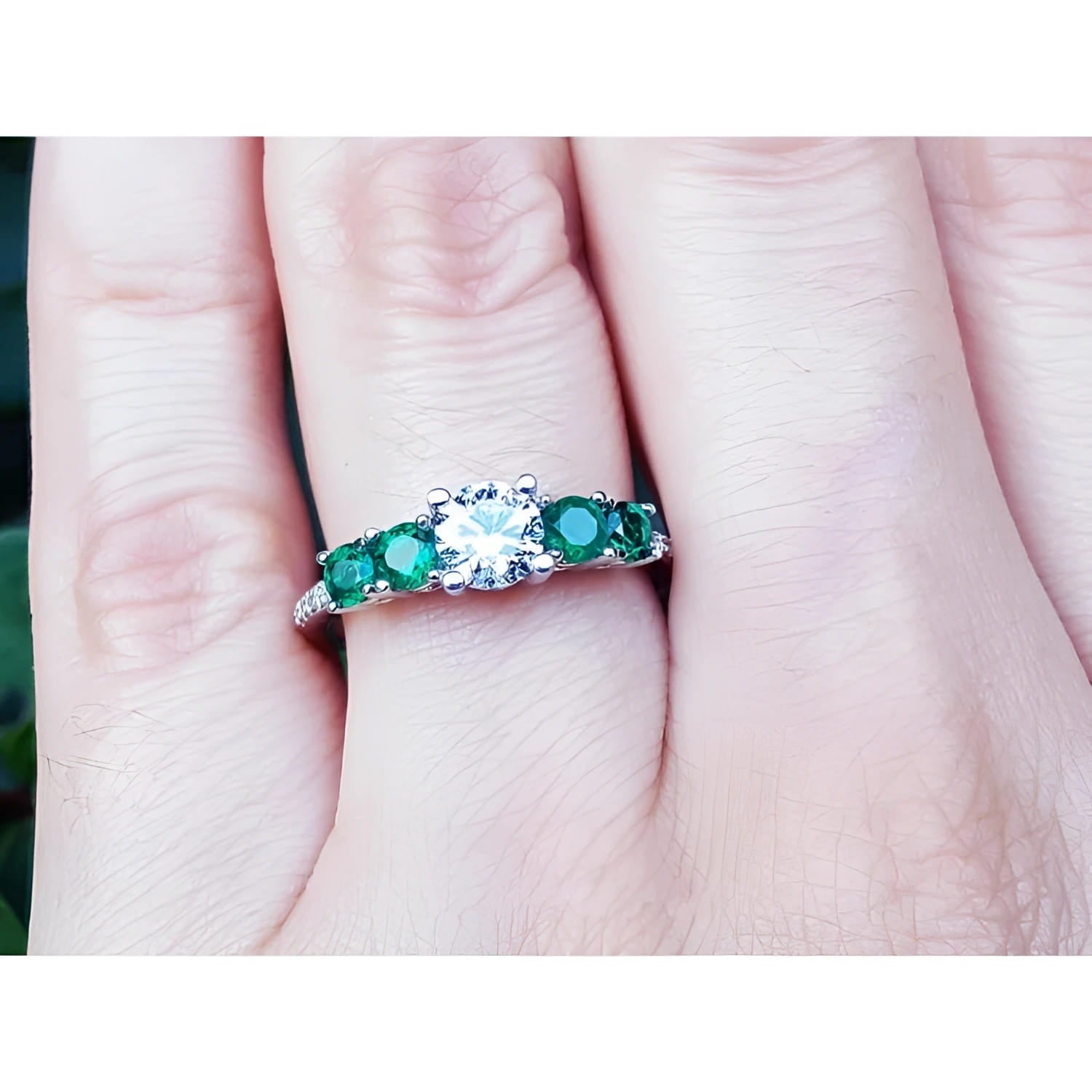 A white gold ring for women with a center round diamond and side round emeralds on a finger.