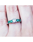 A white gold ring for women with a center round diamond and side round emeralds on a finger.