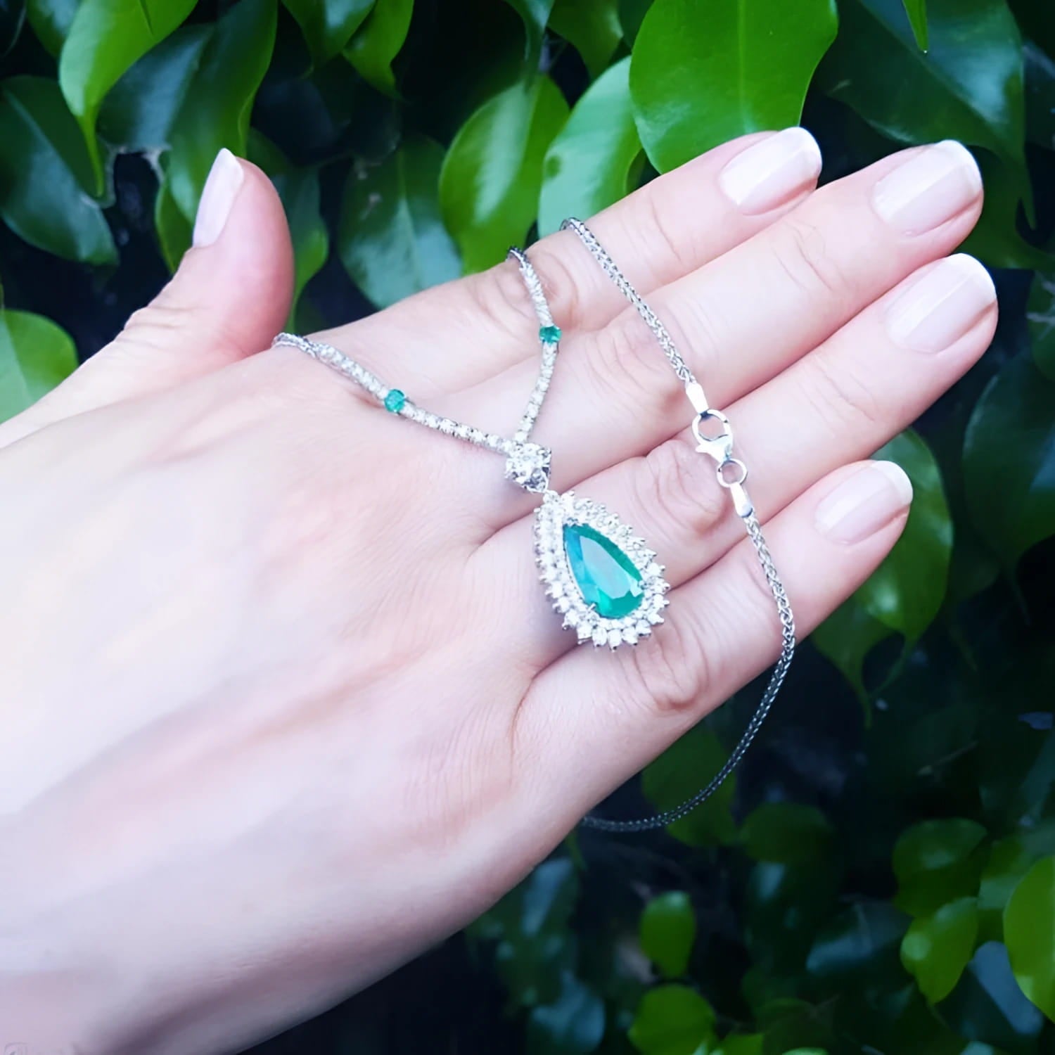 Pear cut emerald necklace in 18K white gold with double diamond halo, held in a hand.
