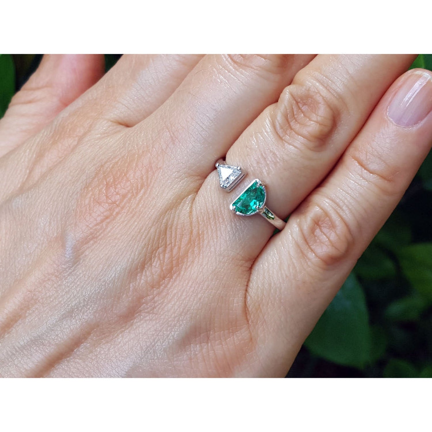 18k white gold emerald and diamond you and me ring, shown worn on a person's finger against a blurred green background.