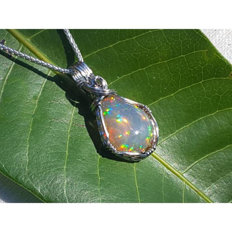 Welo Ethiopian multicolor opal pendant with pinpoint colors, wrapped in silver wire, on a green leaf.
