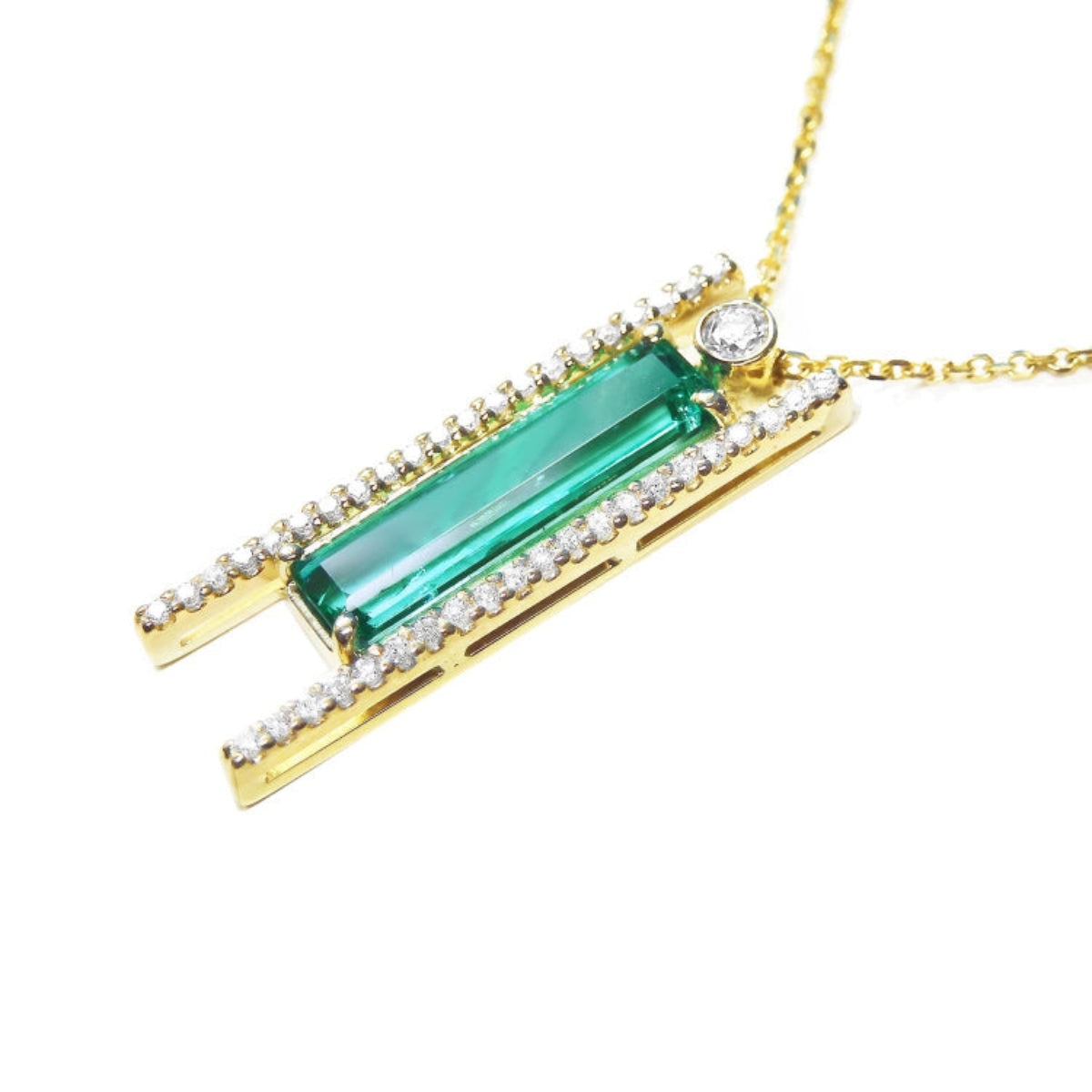 Left side of a gold necklace with a natural baguette cut emerald gemstone and side round diamonds on a white background