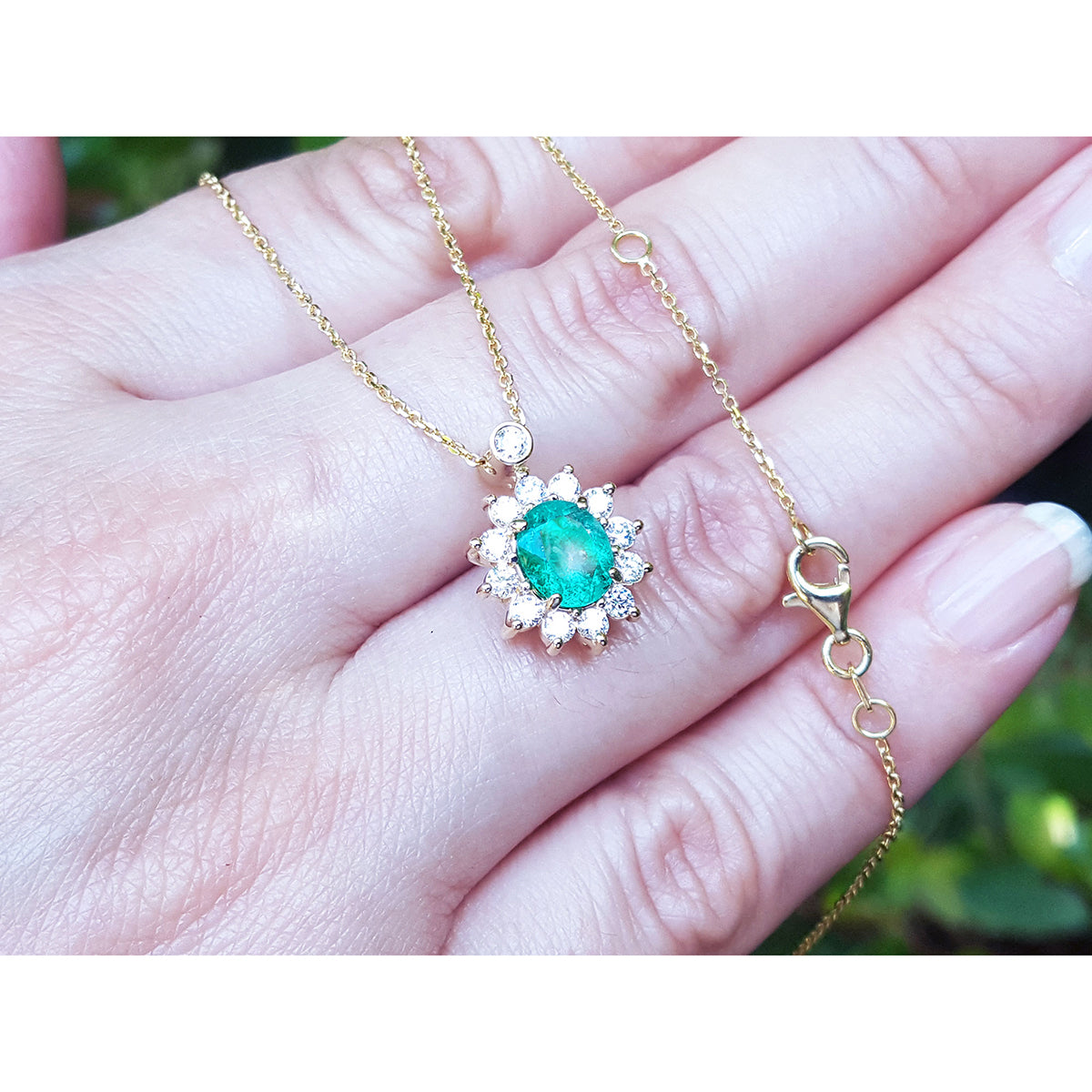 Enlarged view of a gold necklace with an emerald gemstone pendant held in a hand against a blurred natural background