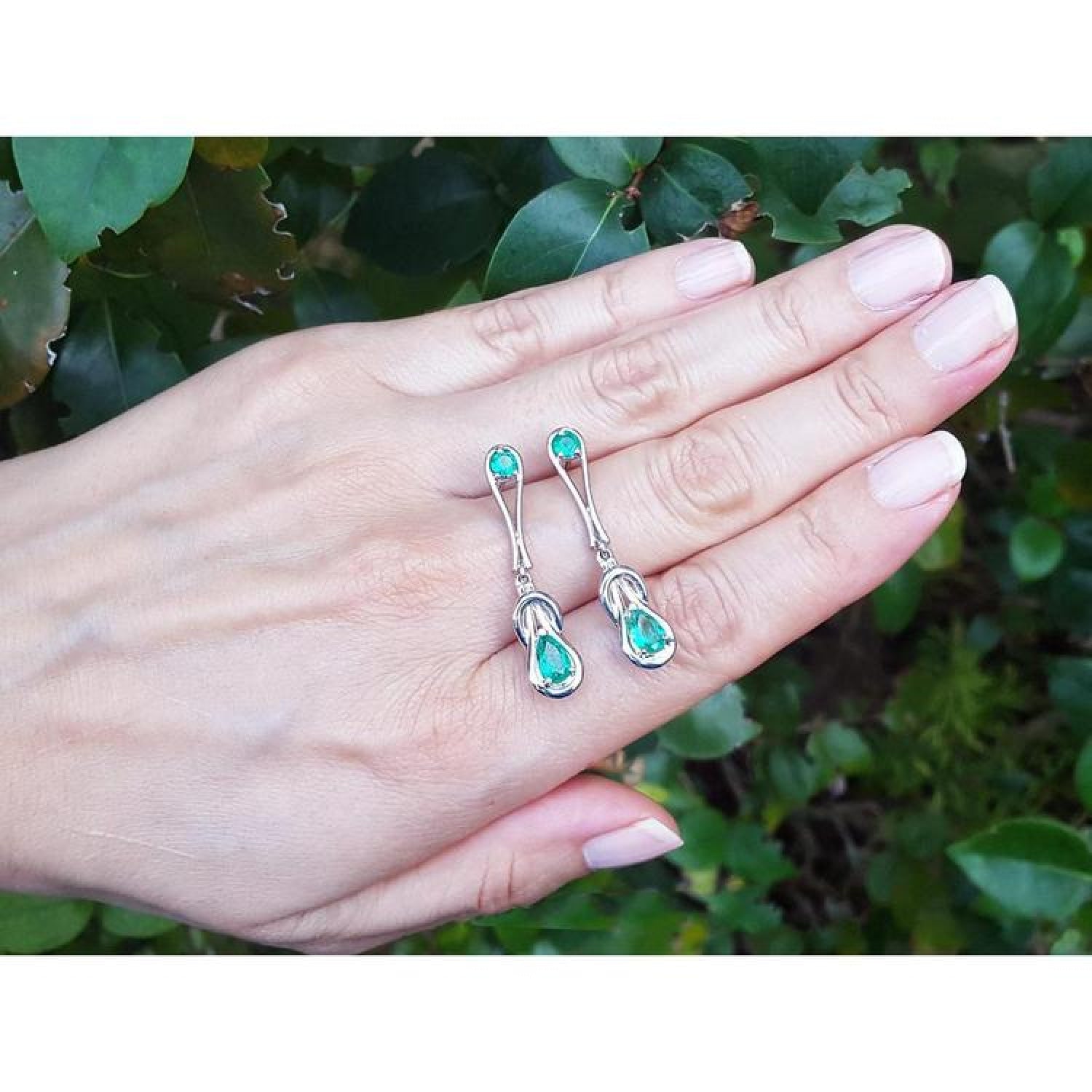 14K white gold love knot earrings with pear-shaped Colombian emeralds, held in a hand.