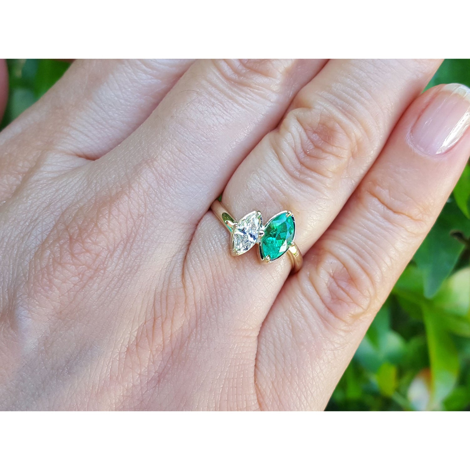 You and me emerald and diamond marquise ring shown worn on a person's finger against a blurred green background.