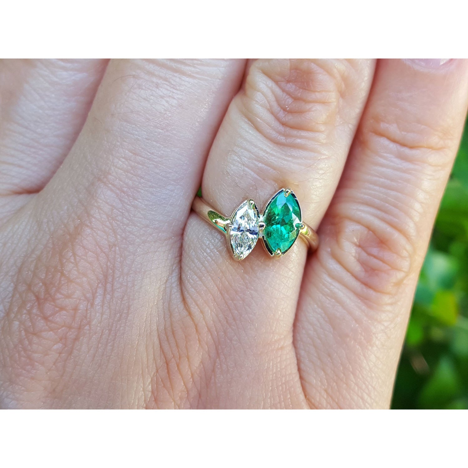 A 18k gold ring with a marquise-cut emerald and a diamond, shown worn on a person's finger against a blurred green background.