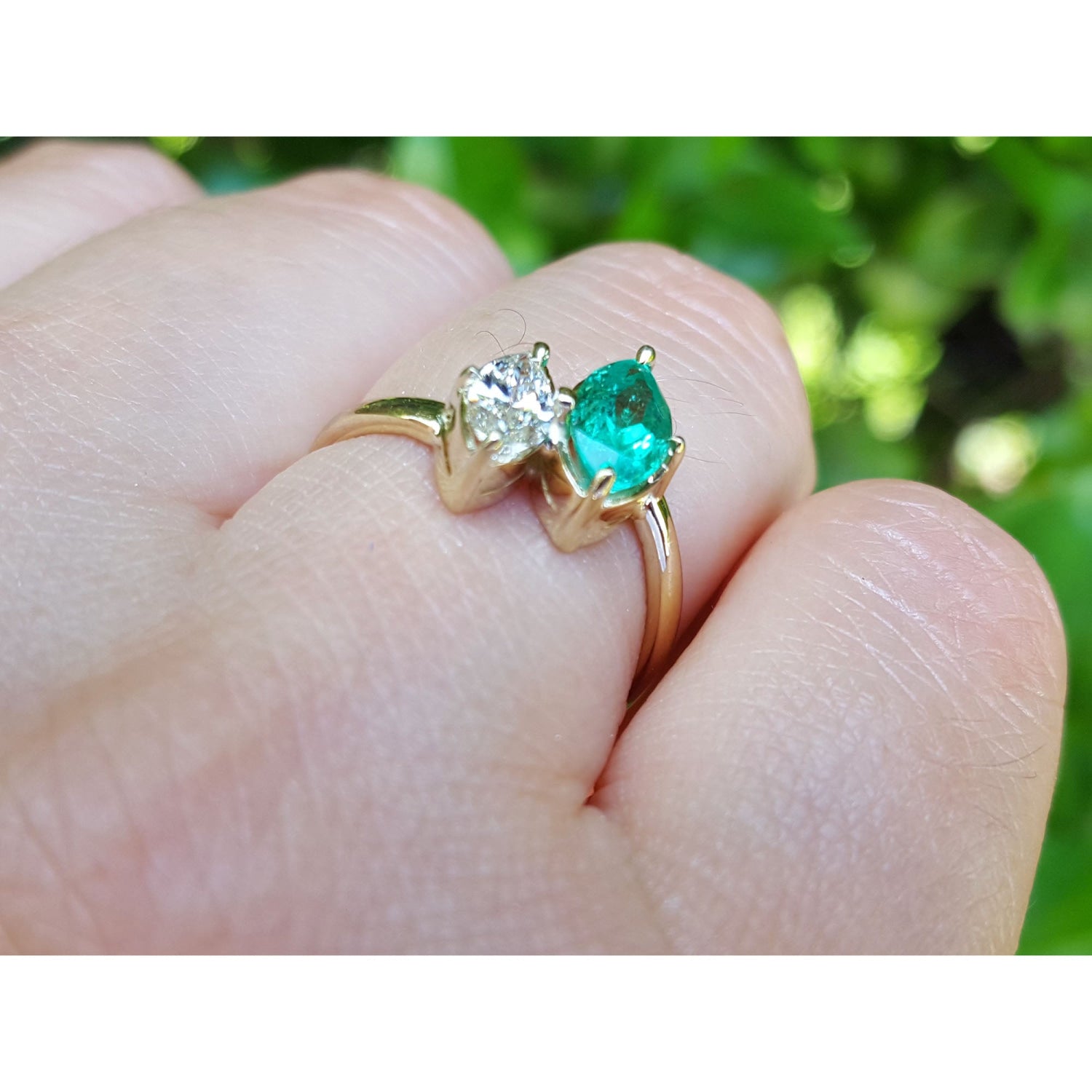 A gold ring with a marquise-cut emerald and diamond, worn on a person's finger with a blurred green outdoor background.