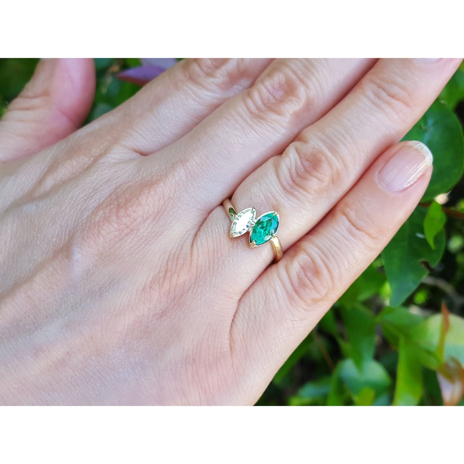 A 18k yellow gold ring toi et moi design with marquise emeralds and diamond shown worn on a person's finger with a blurred green garden background.