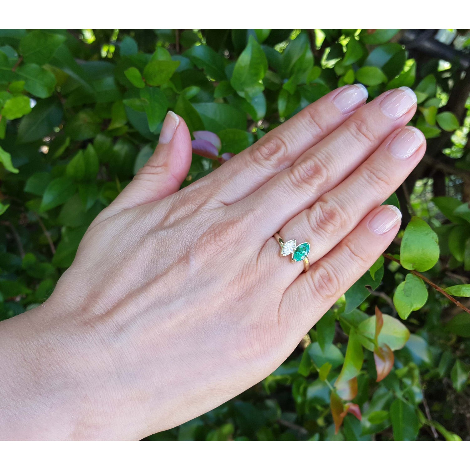 You and me emerald and diamond ring, worn on the ring finger of a hand, set against a background of green outdoor foliage.