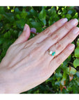 You and me emerald and diamond ring, worn on the ring finger of a hand, set against a background of green outdoor foliage.