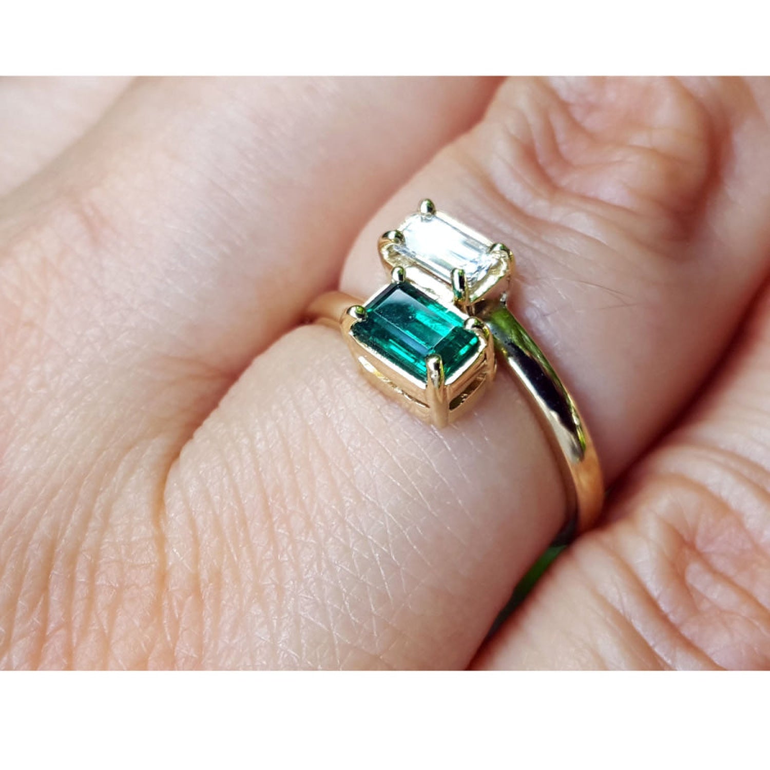 A gold ring with a large emerald-cut green gemstone and a smaller clear rectangular gemstone, shown worn on a person's finger.