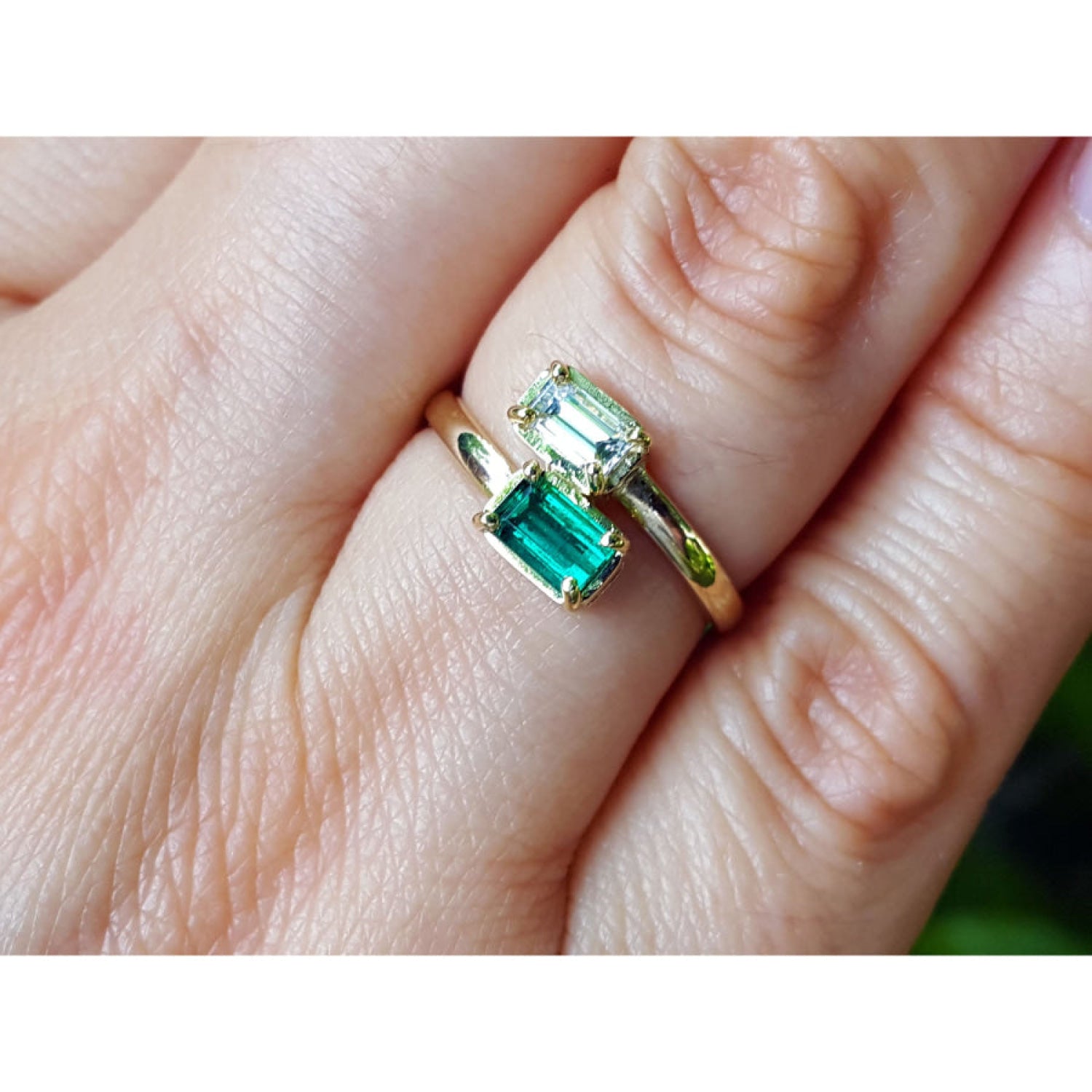 A close-up of a hand wearing a gold ring with two rectangular emerald-cut green gemstones.