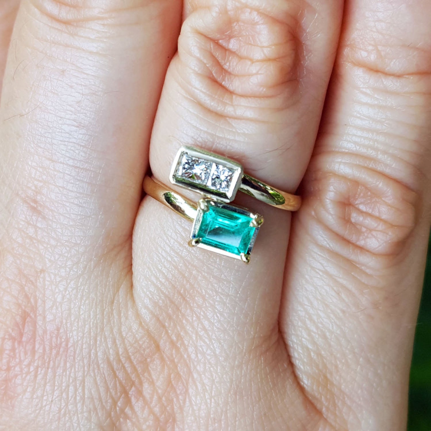 A close-up of a hand wearing a you and me 18k gold ring with a natural emerald rectangular emerald-cut and diamonds.