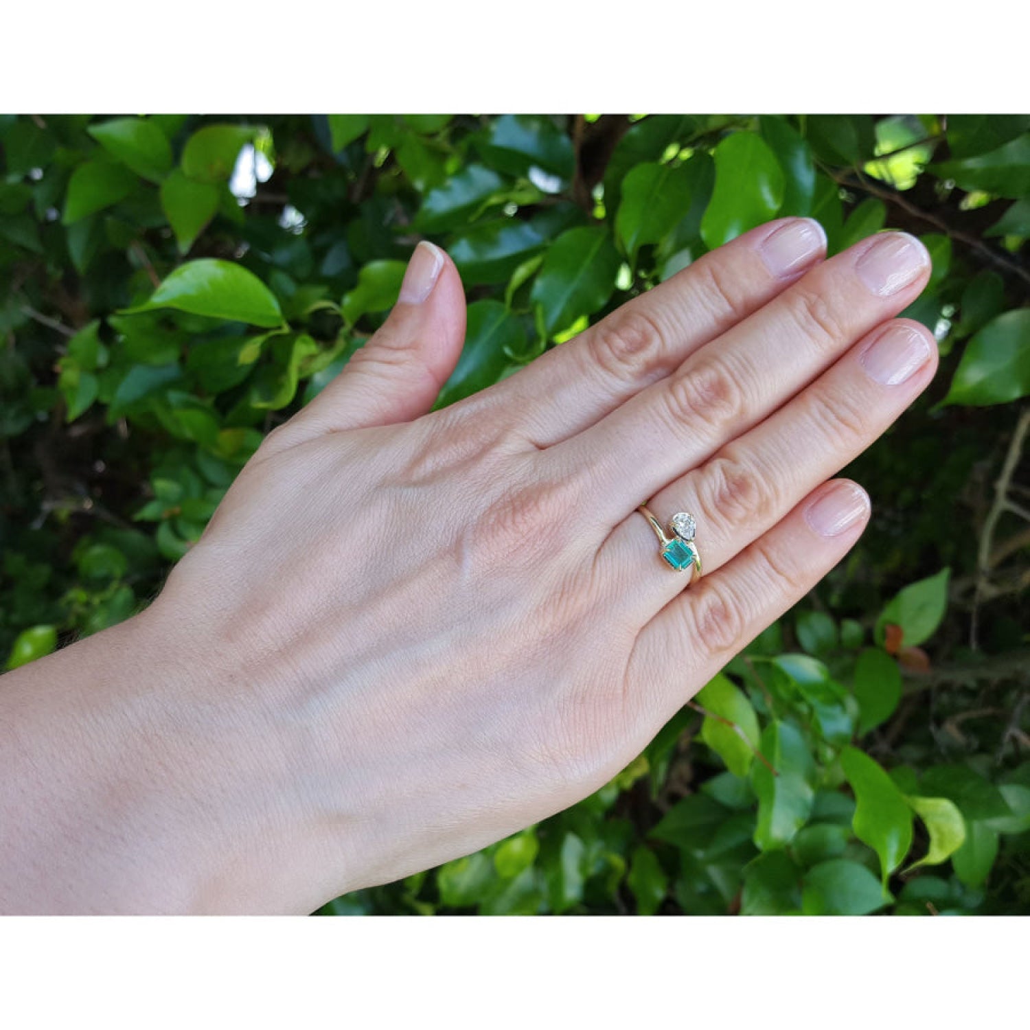 A gold ring toi at moi style with a natural emerald paired with a pear cut diamond worn on a person's ring finger, shown against a background of green foliage.