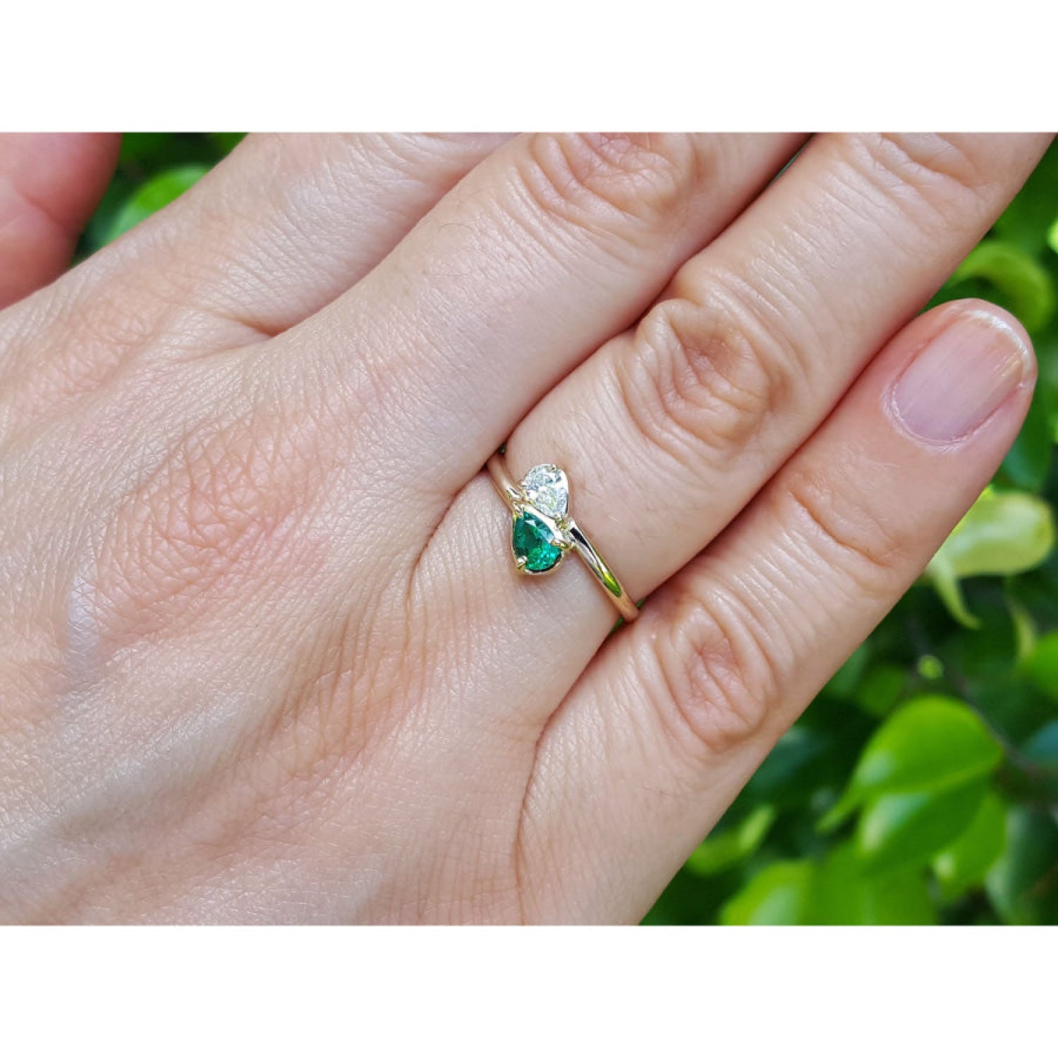 A gold ring with a pear-cut emerald and a diamond, worn on a person's finger with a blurred green outdoor background.