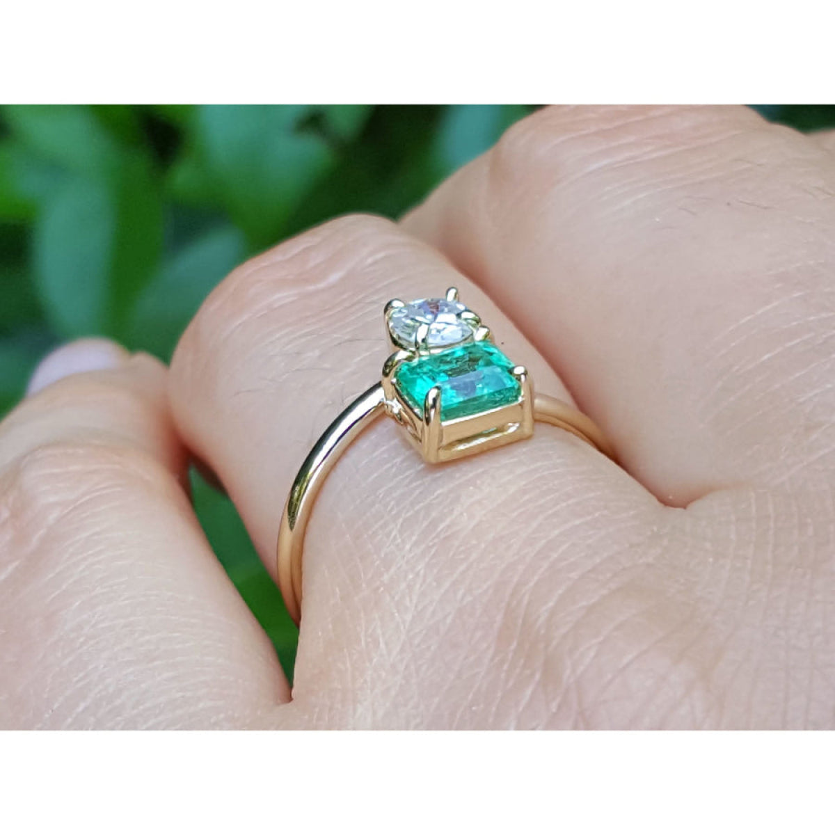 A yellow gold You and Me ring with a natural colombian emerald and diamond, shown worn on a finger against a blurred green background.