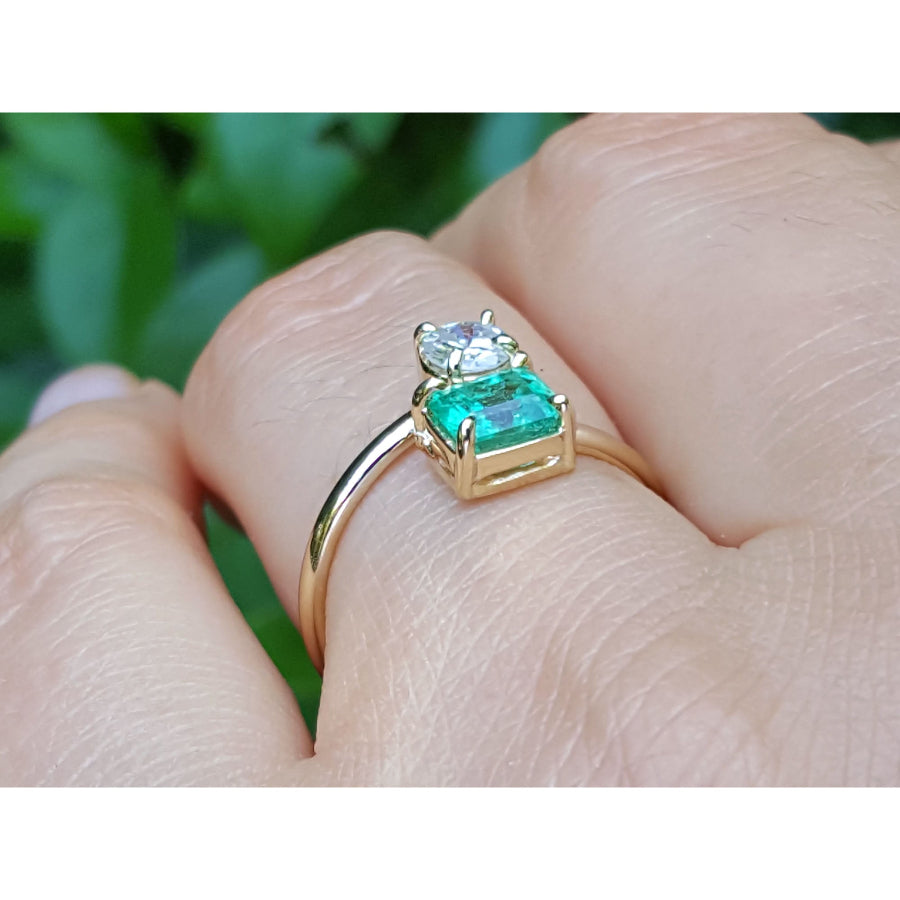 A yellow gold You and Me ring with a natural colombian emerald and diamond, shown worn on a finger against a blurred green background.