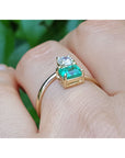 A yellow gold You and Me ring with a natural colombian emerald and diamond, shown worn on a finger against a blurred green background.