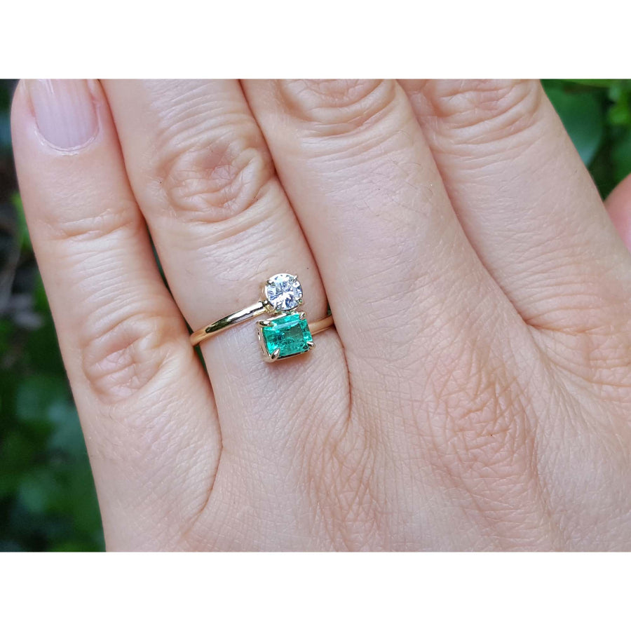 A yellow gold toi et moi emerald and diamond ring, shown worn on a person's finger against a blurred green background.