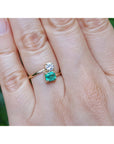 A yellow gold toi et moi emerald and diamond ring, shown worn on a person's finger against a blurred green background.