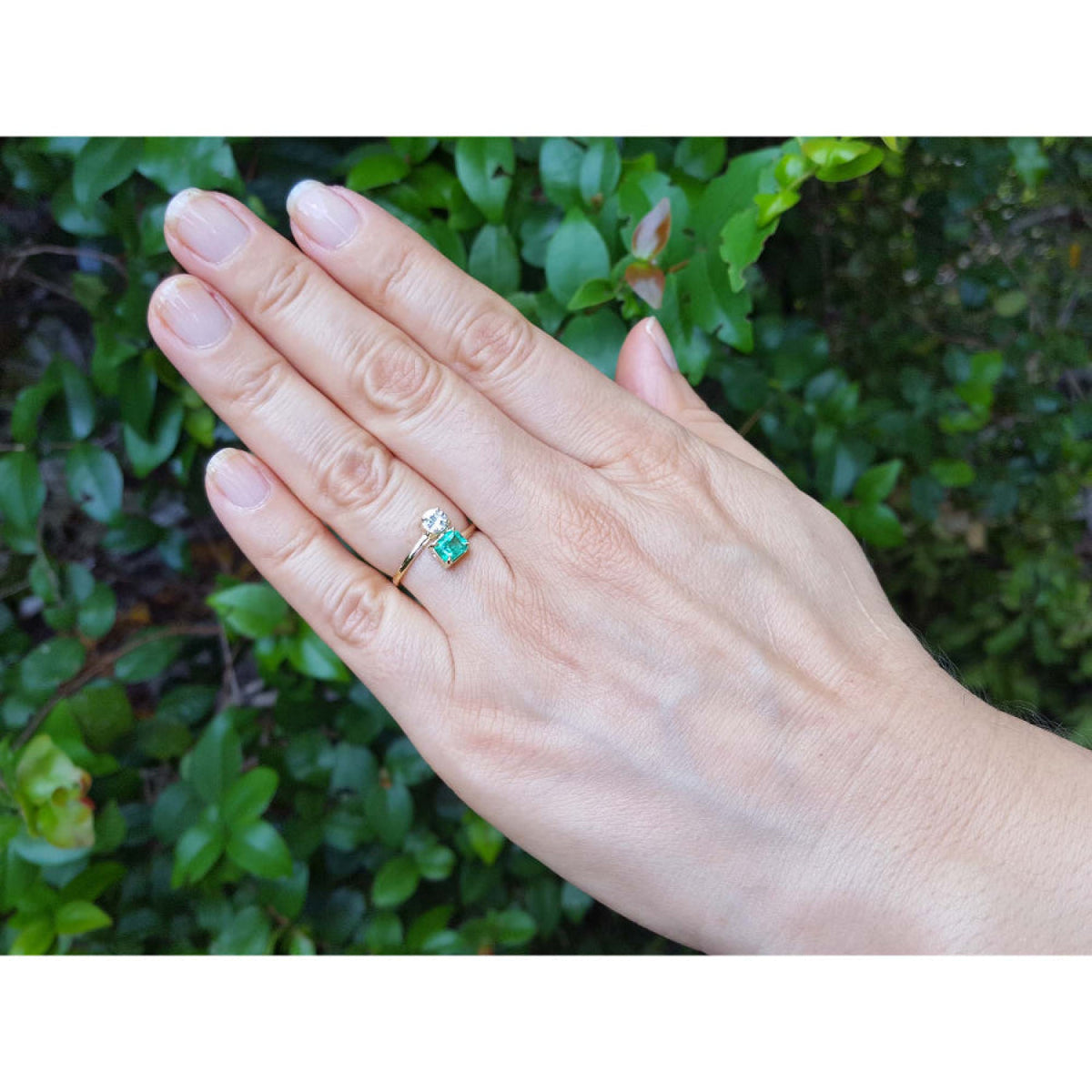 A 18k gold ring with a natural colombian emerald worn on the ring finger of a hand, set against a background of green foliage.
