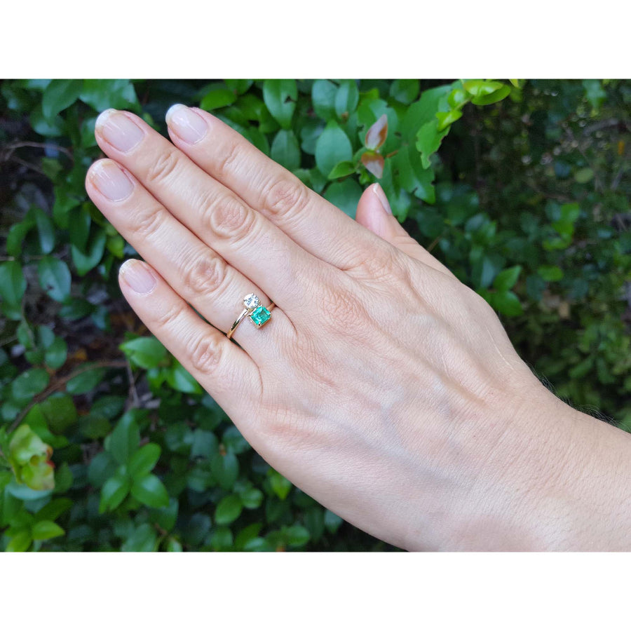 A 18k gold ring with a natural colombian emerald worn on the ring finger of a hand, set against a background of green foliage.