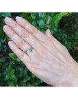 A 18k gold ring with a natural colombian emerald worn on the ring finger of a hand, set against a background of green foliage.