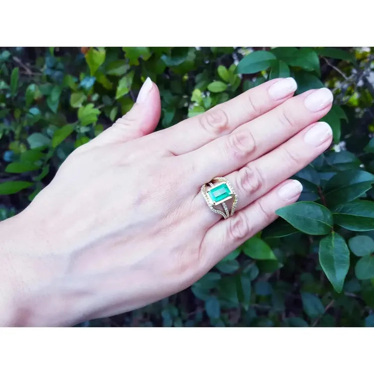 La mano de una mujer luciendo un anillo de oro amarillo con esmeralda sobre un fondo de follaje natural.