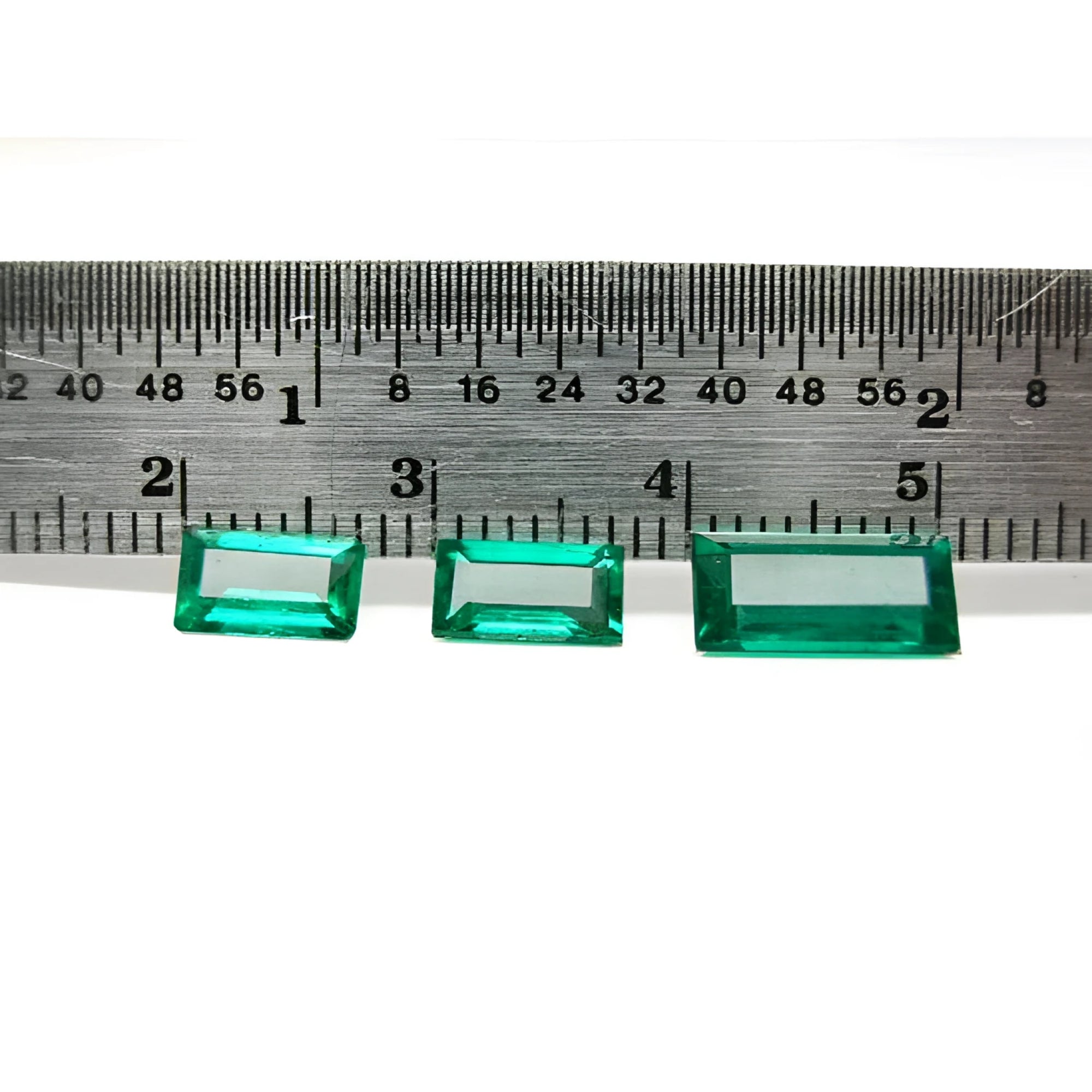 Three baguette-cut natural loose Colombian emeralds, deep bluish-green, displayed with a ruler for scale.