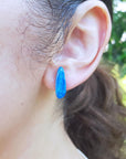 Close-up of an ear wearing a blue opal earring with a blurred green background.
