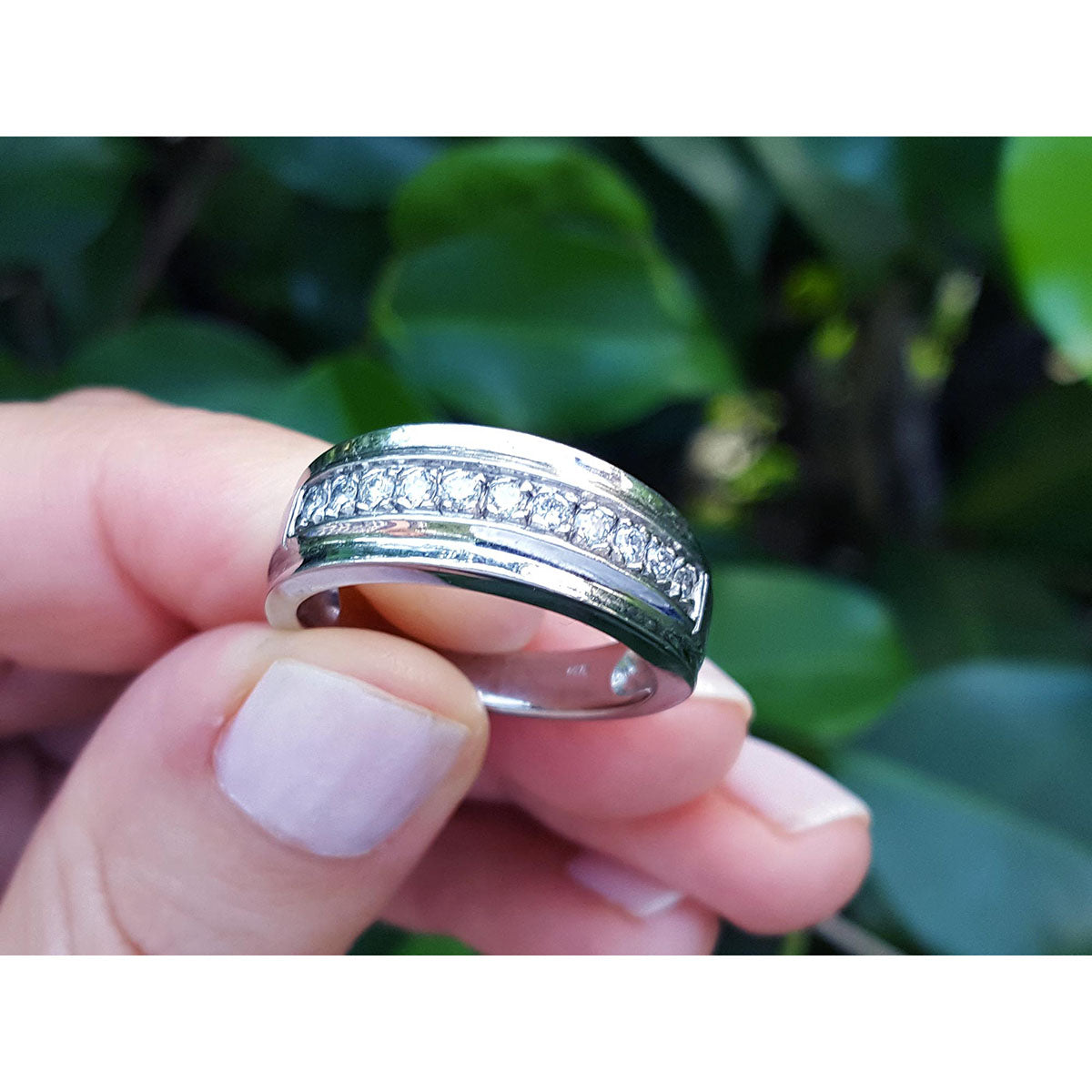 Natural diamond band ring for men in 14K white gold, held by fingers against a blurred green leafy background