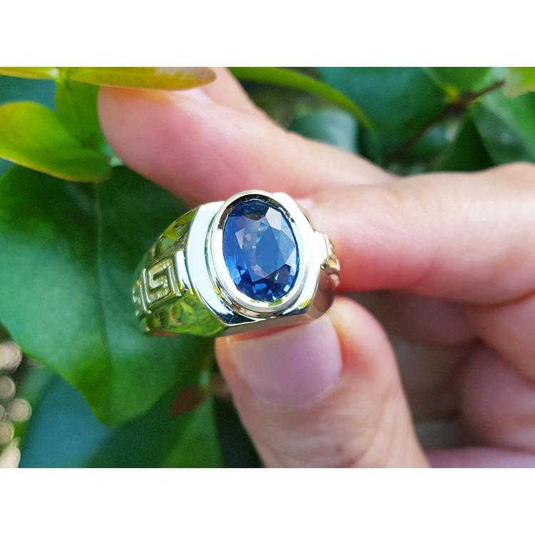 Men's genuine blue sapphire solitaire ring oval cut, top view held between fingers with leafy background