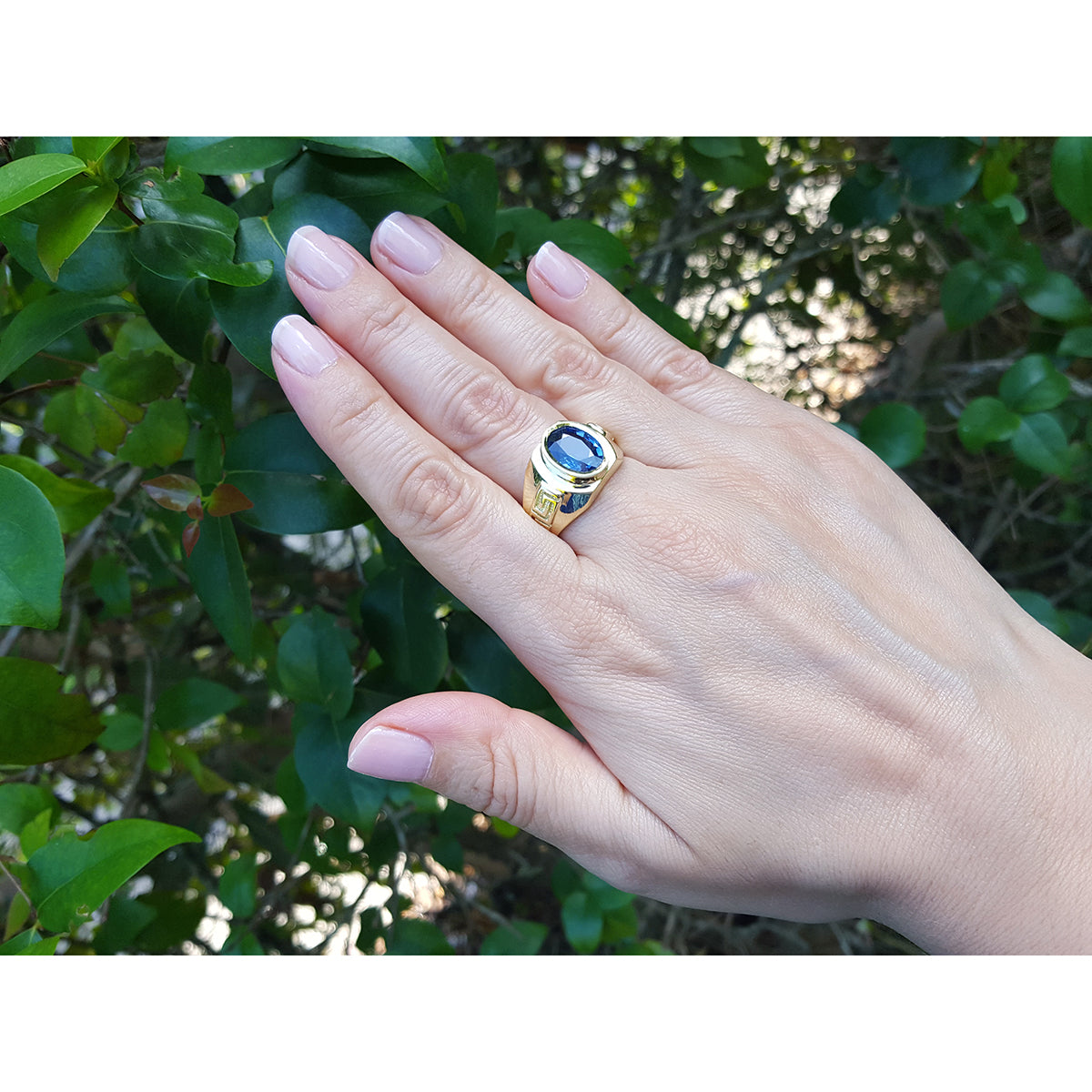 Hand wearing a gold blue sapphire ring against a green leafy background