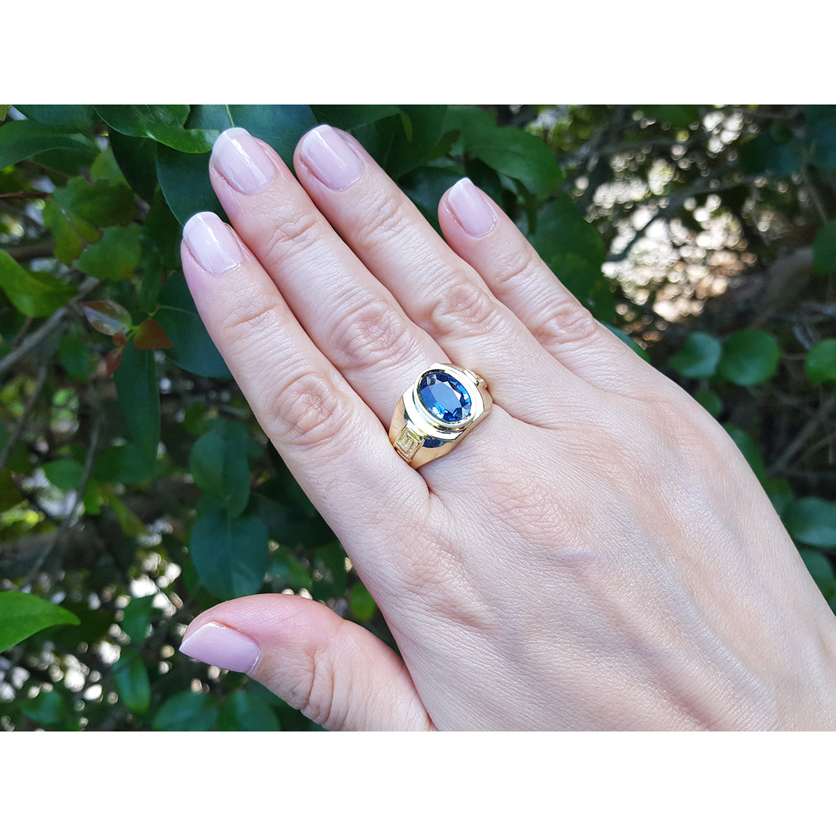 Hand wearing a gold blue sapphire solitaire ring with a blue gemstone against a green leafy background