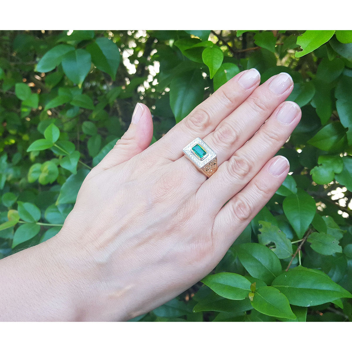 Hand wearing a ring for men with a genuine emerald against a leafy background.