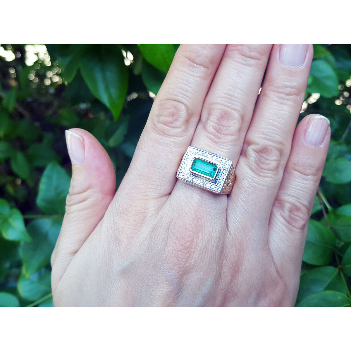 Hand wearing a men&#39;s gold ring with a colombian emerald against a leafy background.