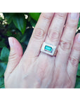 Hand wearing a men's gold ring with a colombian emerald against a leafy background.