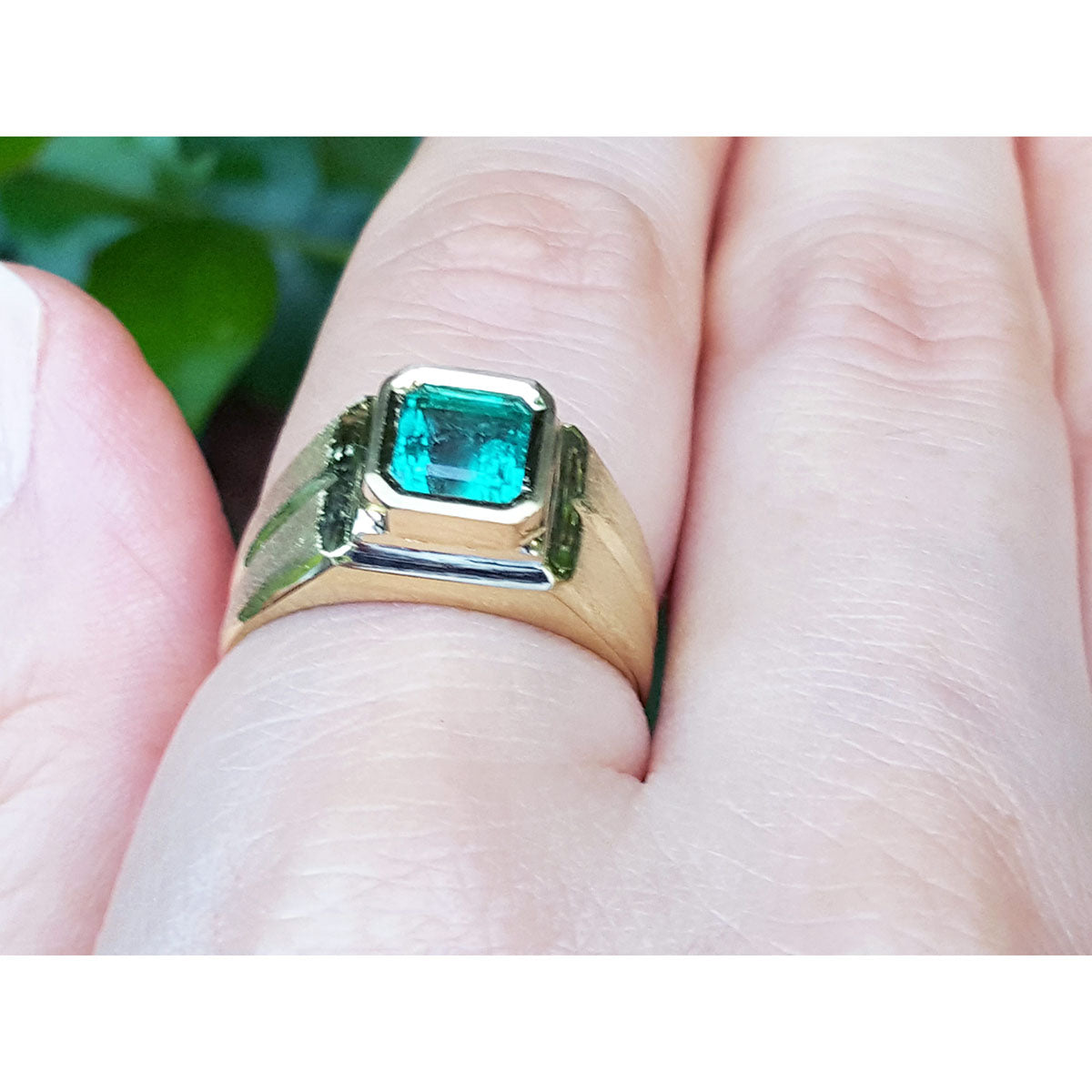 Gold ring for man with a bezel set emerald e on a finger against a blurred natural background.