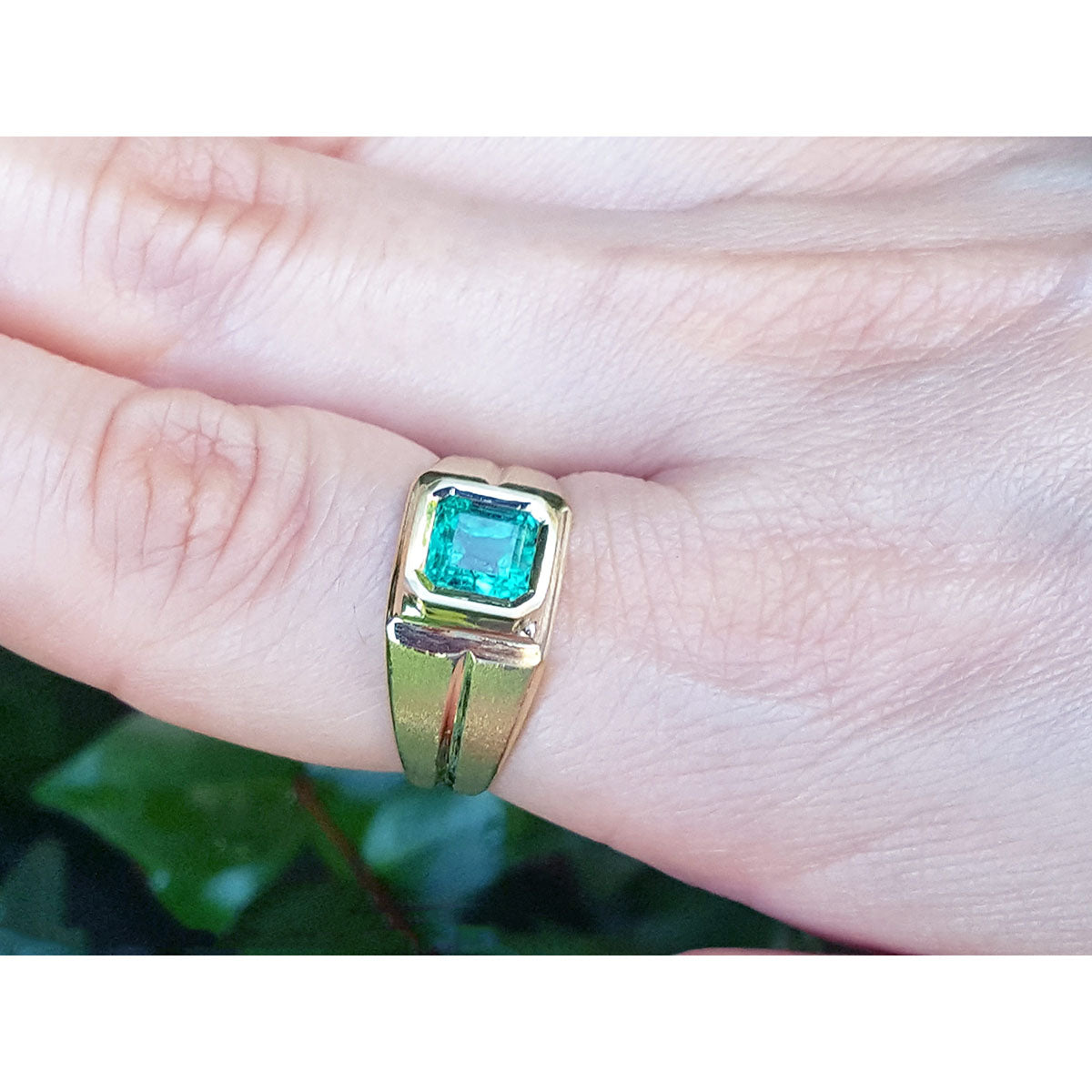 A yellow gold mens emerald pinky ring on a finger against a blurred natural background.