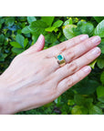 Hand wearing a gold ring with a natural colombian emerald against a green leafy background.