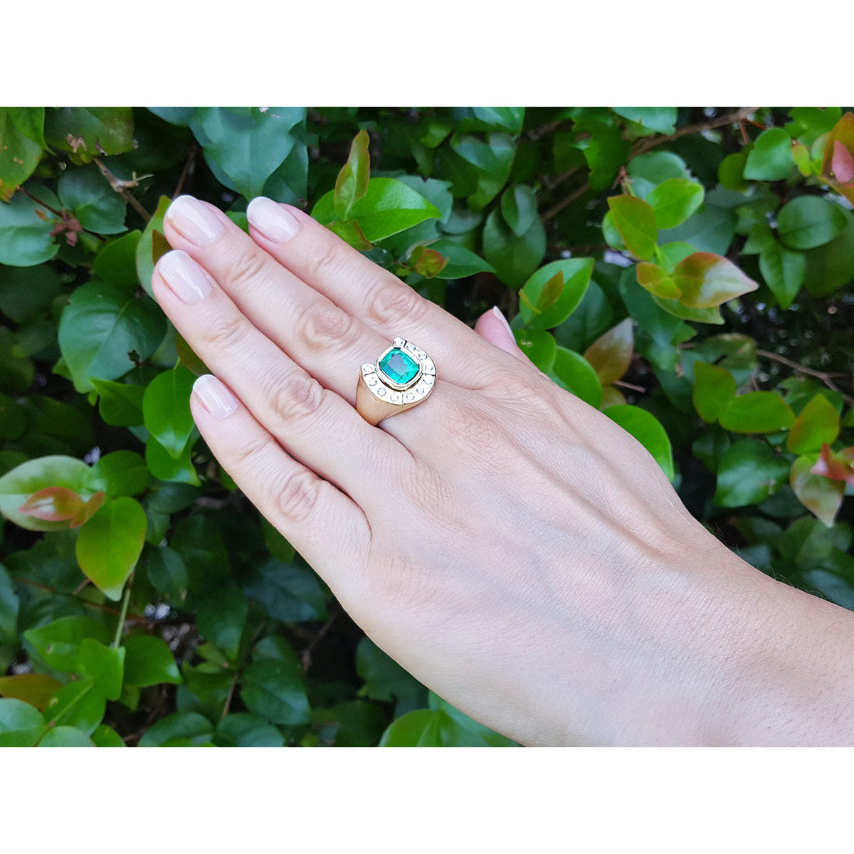 Hand wearing a mens horseshoe ring with a natural colombian emerald against a green leafy background.
