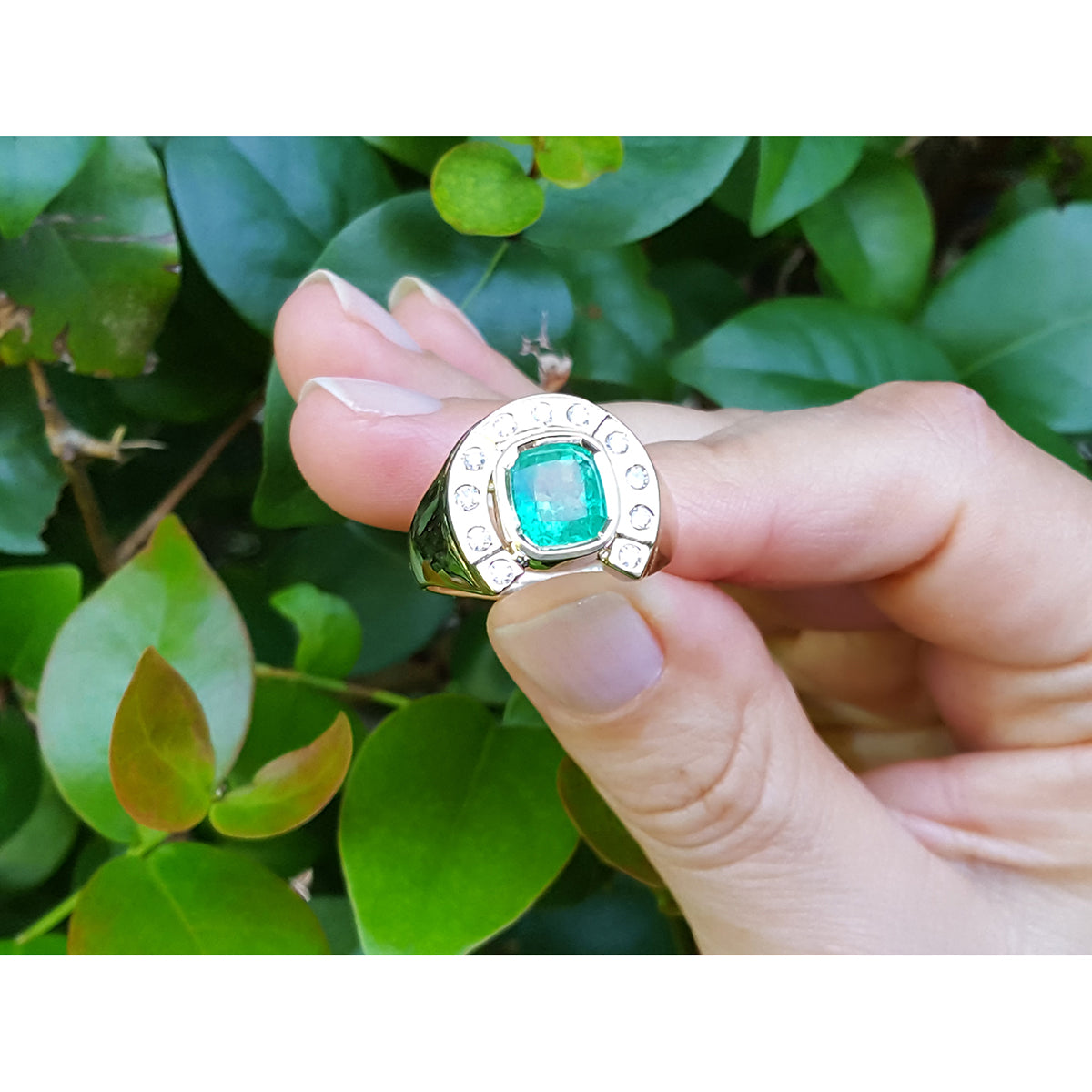 Gold horseshoe ring design with a colombian emerald held by a hand against a green leafy background.