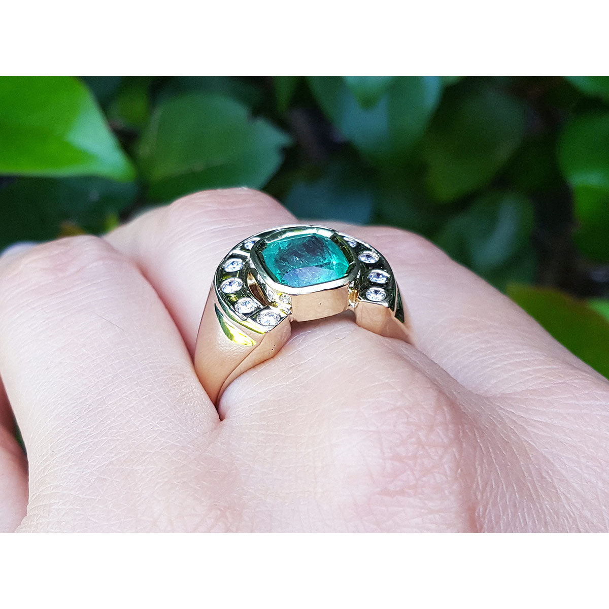 Gold ring with a natural emerald on a hand against a blurred green leafy background.