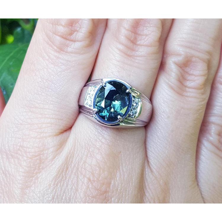 White ring with a blue natural sapphire oval cut on a finger against a natural background