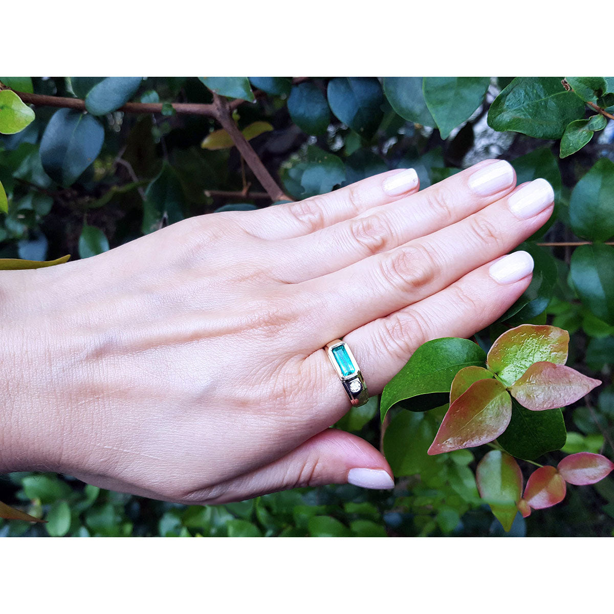 A hand wearing a men&#39;s yellow gold emerald ring against a green leafy background.