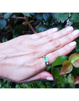 A hand wearing a men's yellow gold emerald ring against a green leafy background.